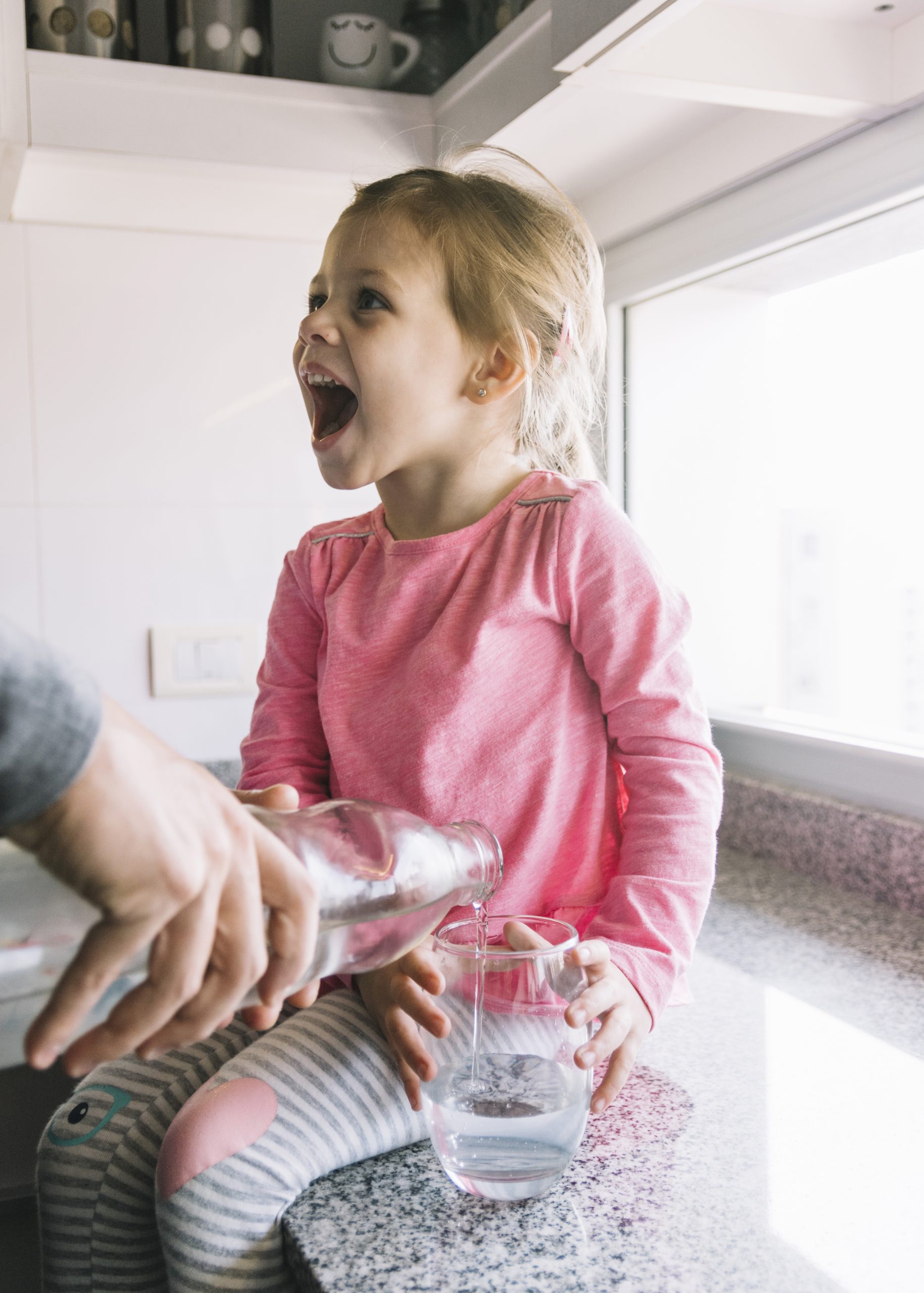 person-s-hand-pouring-water-glass-while-girl-sitting-kitchen-counter